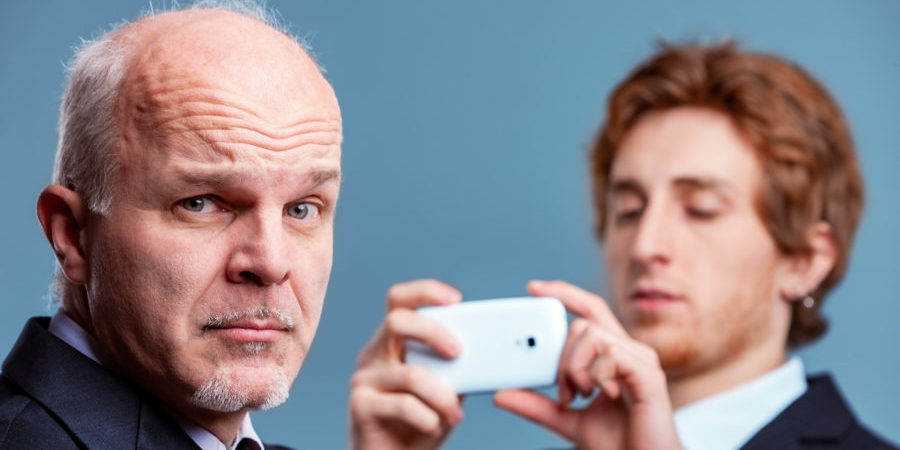 Charismatic conservative senior businessman pulling a wry quizzical face at the camera as a younger colleague or partner prepares to photograph him on his mobile phone